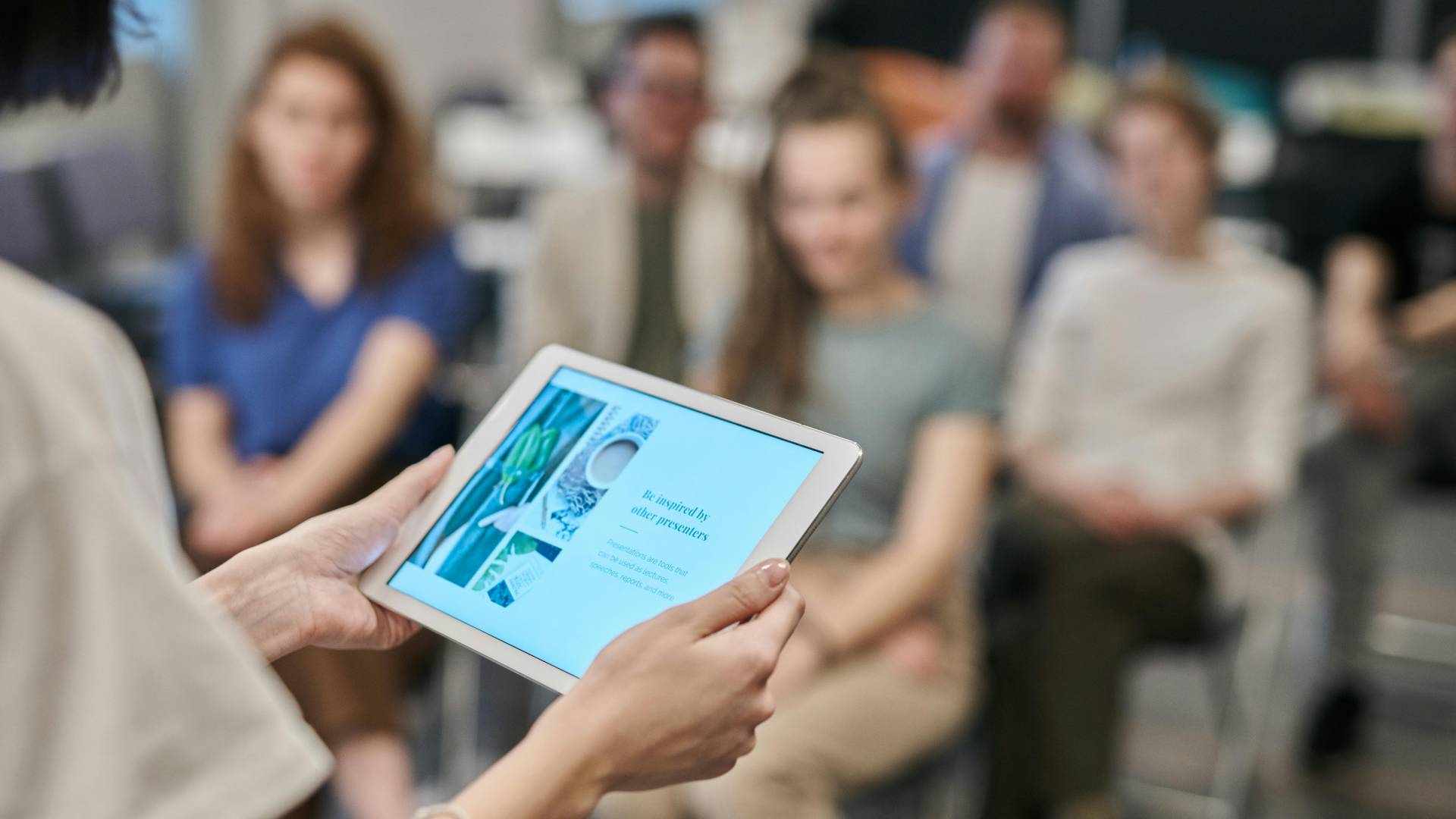 Des mains tiennent une tablette devant des personnes assises qui suivent probablement une formation.
