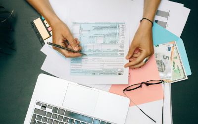 Une personne remplissant un formulaire fiscal (déclaration) sur un bureau, à côté d'un ordinateur portable et de billets de banque.