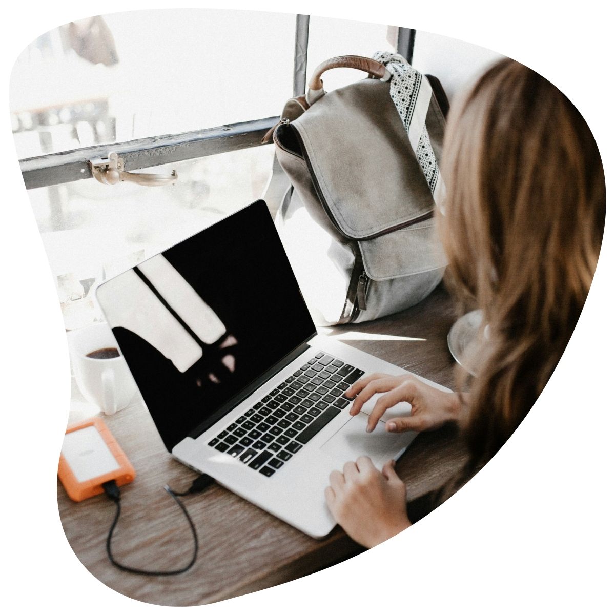 Vue de dos d'une personne travaillant sur un ordinateur portable, avec une tasse de café, un disque dur externe et un sac à dos.