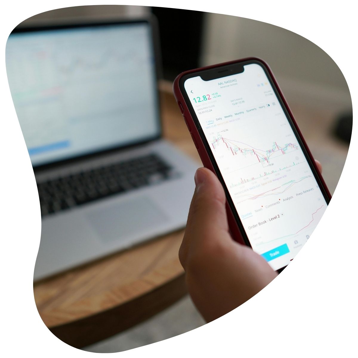 Une main tenant un smartphone affichant un graphique financier, avec un ordinateur portable en arrière-plan.