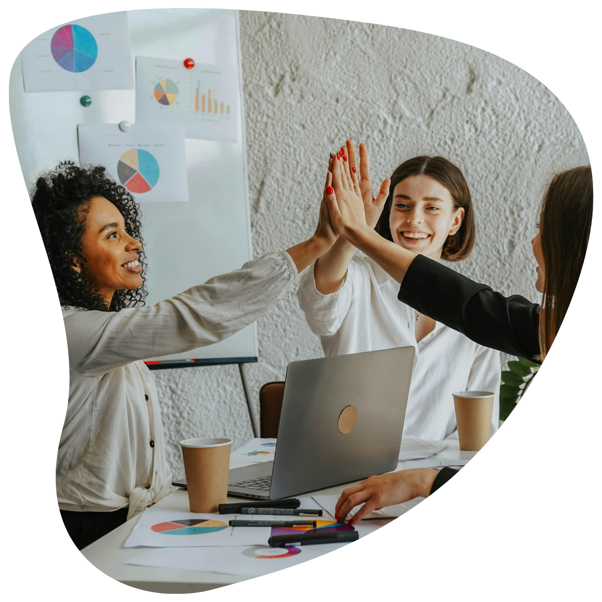 Trois femmes souriantes se tapant dans les mains ("high five") autour d'un bureau, avec des graphiques en arrière-plan.