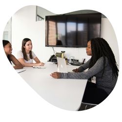 Trois femmes assises autour d'une table de réunion discutant devant un grand écra