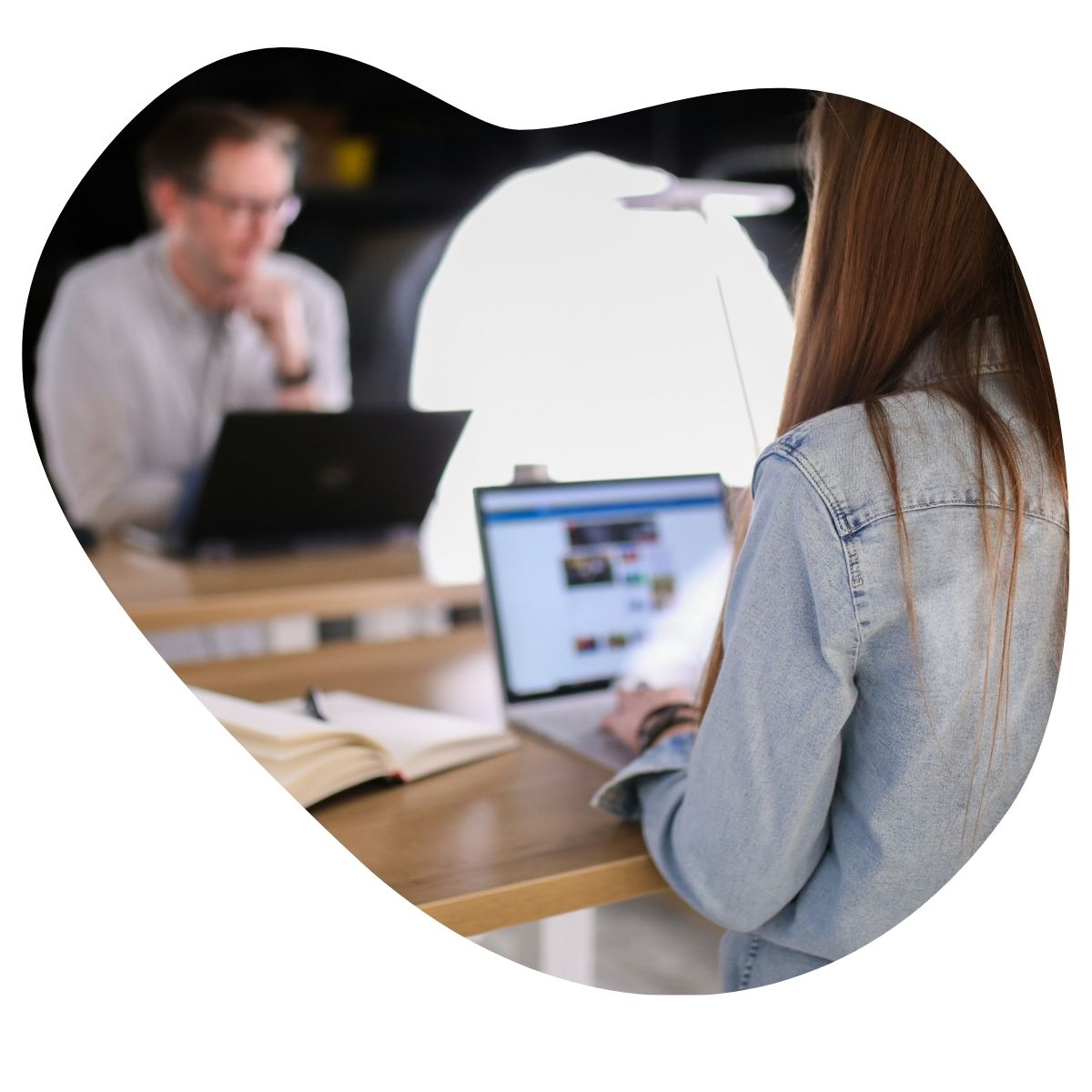 Vue de dos d'une personne travaillant sur un ordinateur portable, avec un collègue en arrière-plan.