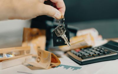 ne main tient une clé attachée à un petit porte-clés en forme de maison. Des billets de 50 euros et une calculatrice sont flous en arrière-plan.