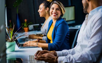 Une professionnelle souriante portant une veste bleue et un haut jaune est assise à son bureau devant un ordinateur portable. Deux collègues sont en arrière-plan.