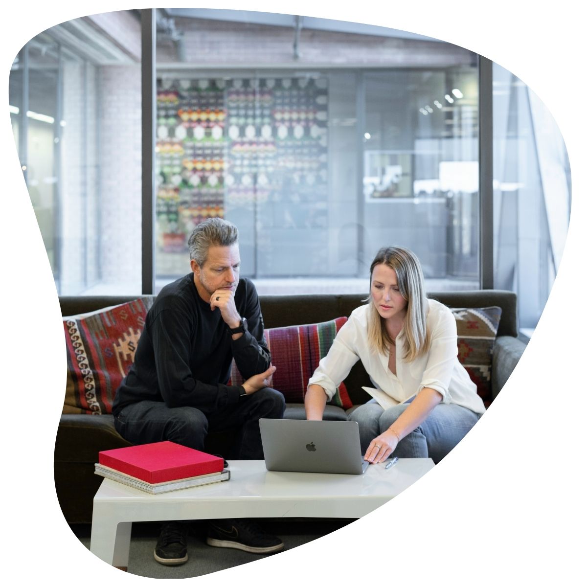 Un homme et une femme sont assis sur un canapé dans un espace de bureau moderne, regardant ensemble un ordinateur portable posé sur une table basse. L'homme est vêtu de noir et la femme d'une chemise blanche. L'image est encadrée par une forme ovale.