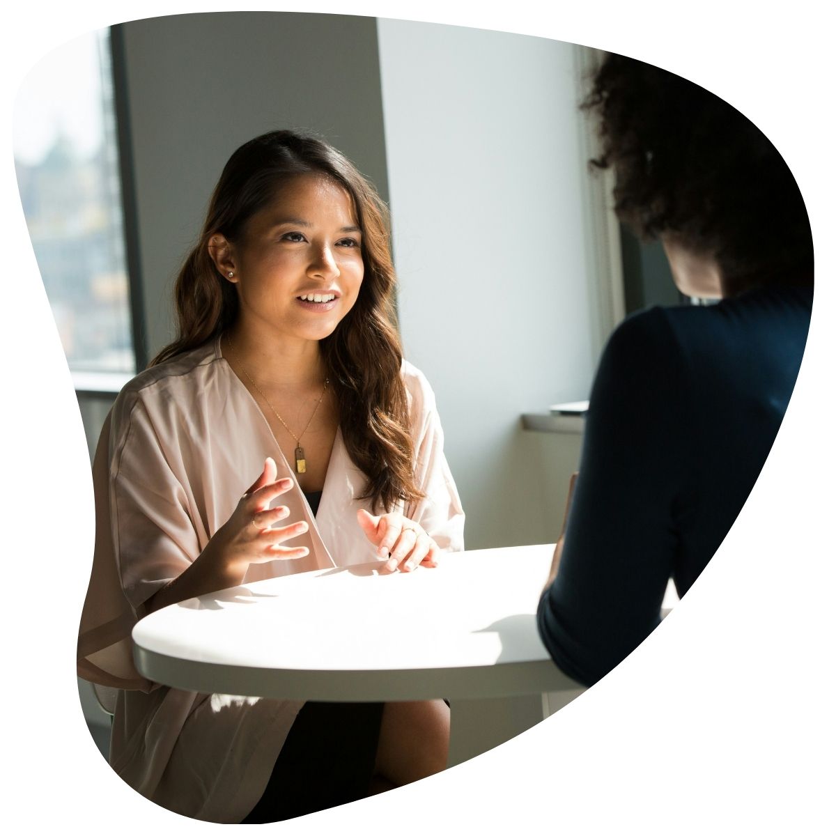 Une jeune femme souriante et aux cheveux bruns, vêtue d'un chemisier clair, est assise à une table ronde blanche et parle à une autre personne. L'image est encadrée par une forme ovale.