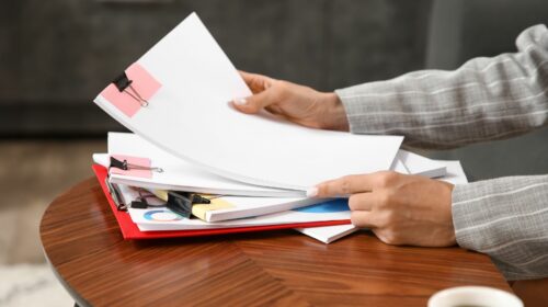Une personne en veste grise manipule une pile de documents et de dossiers imprimés posés sur une table en bois. Les feuilles blanches et les rapports contenant des graphiques en couleurs sont assemblés à l'aide de pince-notes.
