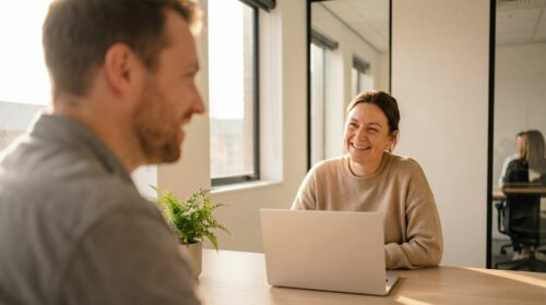 Une femme souriante, en pull beige, discute chaleureusement avec un homme (vu de trois-quarts dos et flou au premier plan) lors d'un entretien ou d'une réunion de travail dans un bureau lumineux