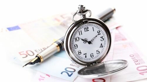 Une montre à gousset en argent ouverte, laissant apparaître son cadran, est posée au premier plan à côté d'un élégant stylo-plume noir et doré[cite: 1]. En arrière-plan, plusieurs billets de banque en euros (notamment de 50, 20 et 10 euros) sont éparpillés sur une surface blanche.